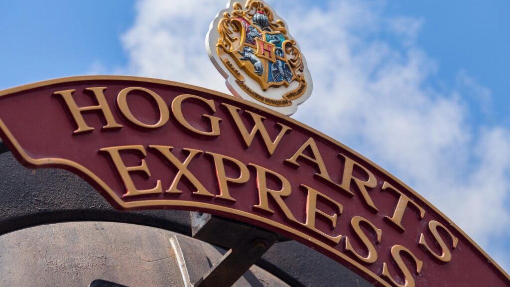 A red and gold "Hogwarts Express" sign with a crest on top is shown against a blue sky with clouds.