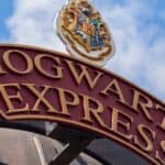 A red and gold "Hogwarts Express" sign with a crest on top is shown against a blue sky with clouds.