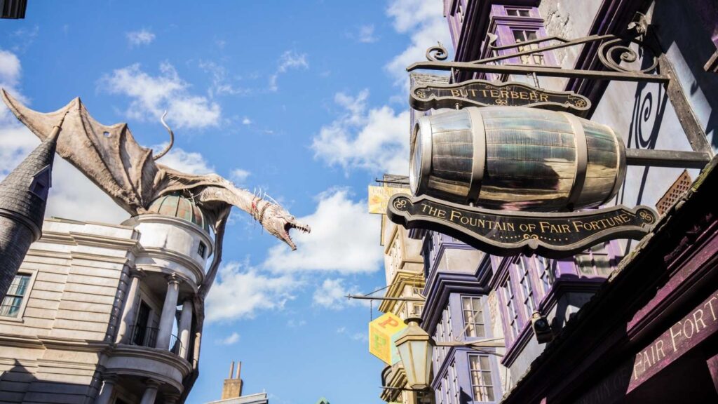 A large dragon statue perches atop a building beside a sign shaped like a barrel reading "Butterbeer, The Fountain of Fair Fortune" on a themed street under a blue sky.