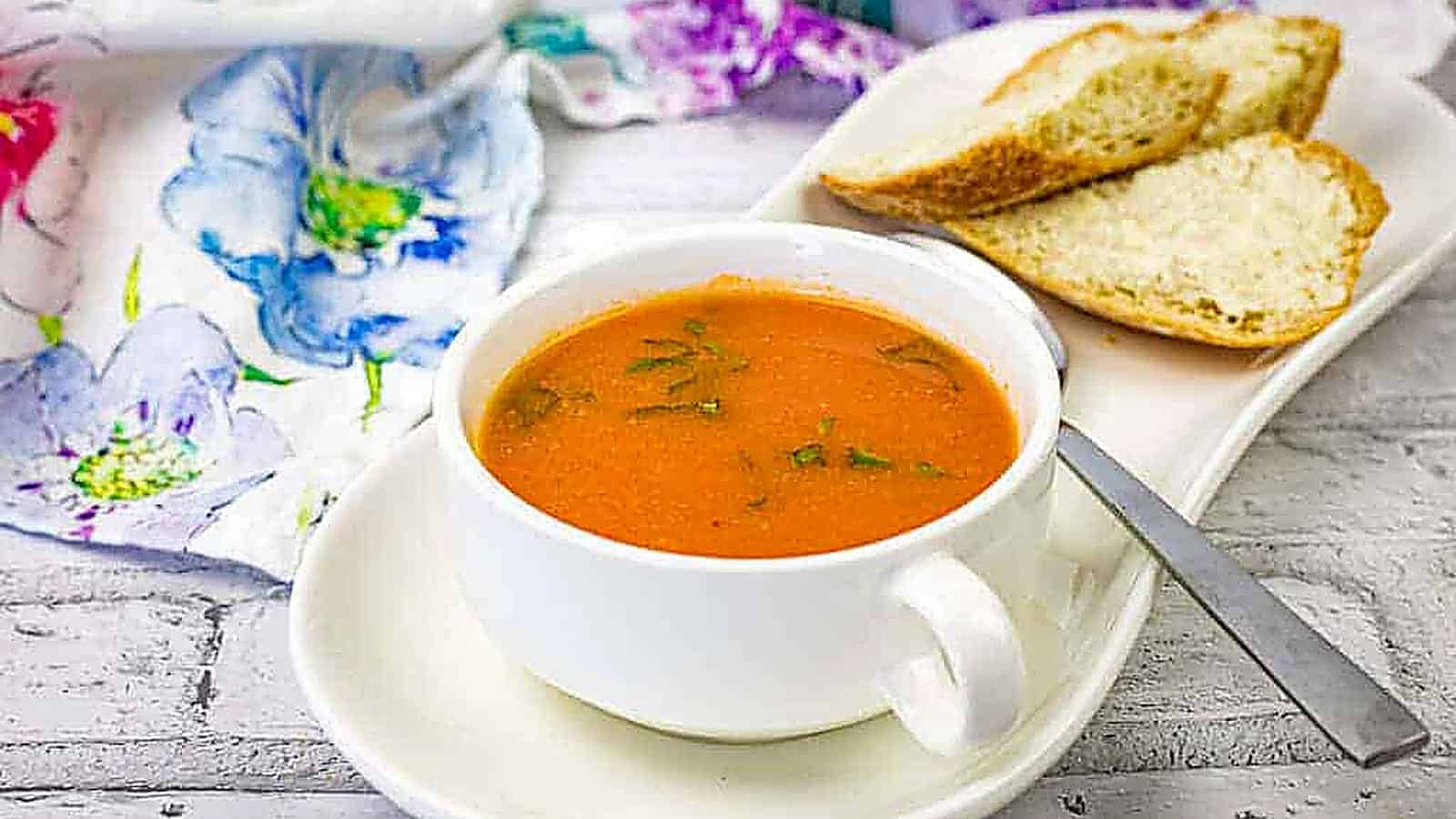 A bowl of old-fashioned tomato soup garnished with herbs, served with slices of crusty bread on a white plate.