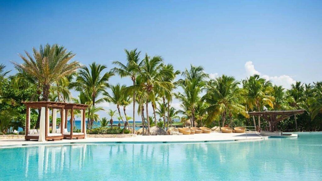 Resort swimming pool with clear blue water, surrounded by palm trees, lounge chairs, and wooden cabanas under a sunny sky.