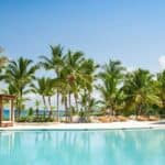 Resort swimming pool with clear blue water, surrounded by palm trees, lounge chairs, and wooden cabanas under a sunny sky.