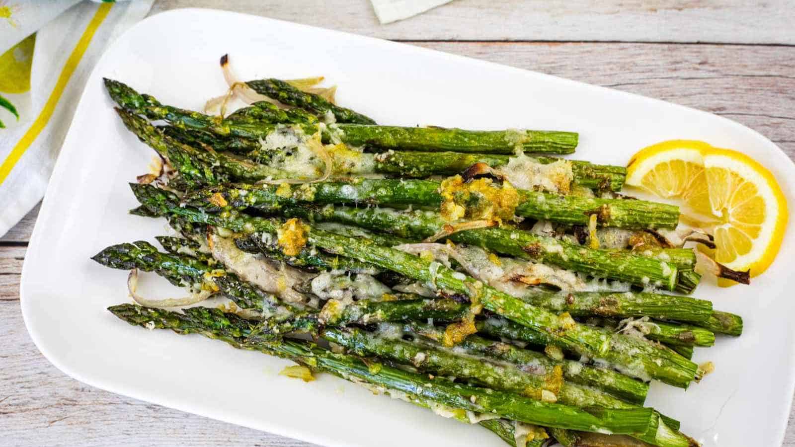 Roasted asparagus spears topped with melted cheese and grated lemon zest, served on a white plate with two lemon slices.