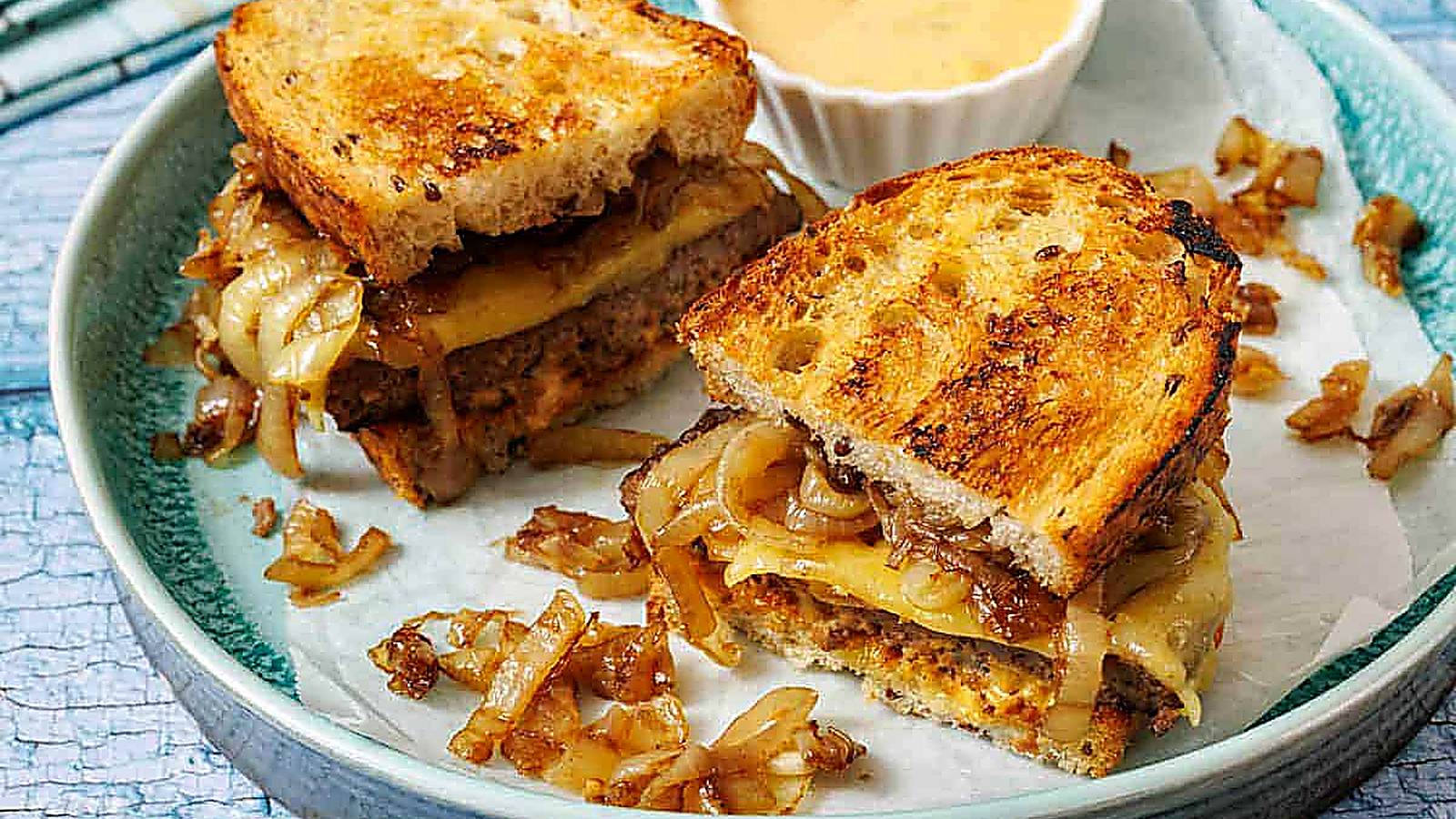 A plate with a Classic Patty Melt cut in half, served with a cup of dipping sauce.