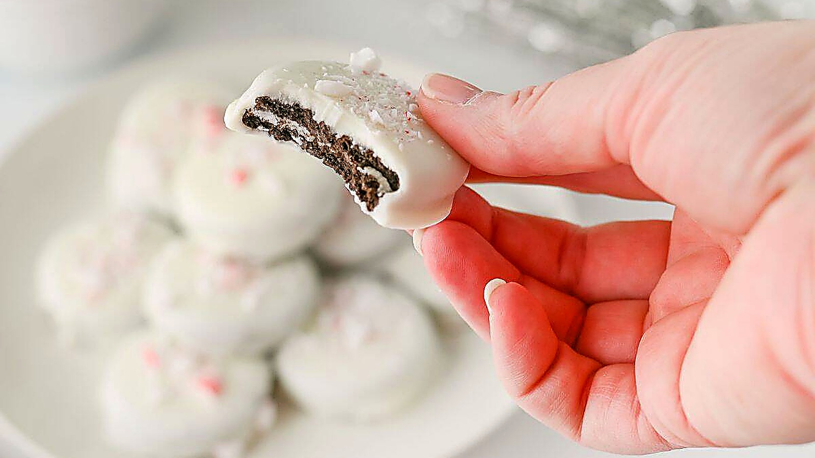 A hand holds a Peppermint Crunch Oreo with a bite taken out, with more cookies on a plate in the background.
