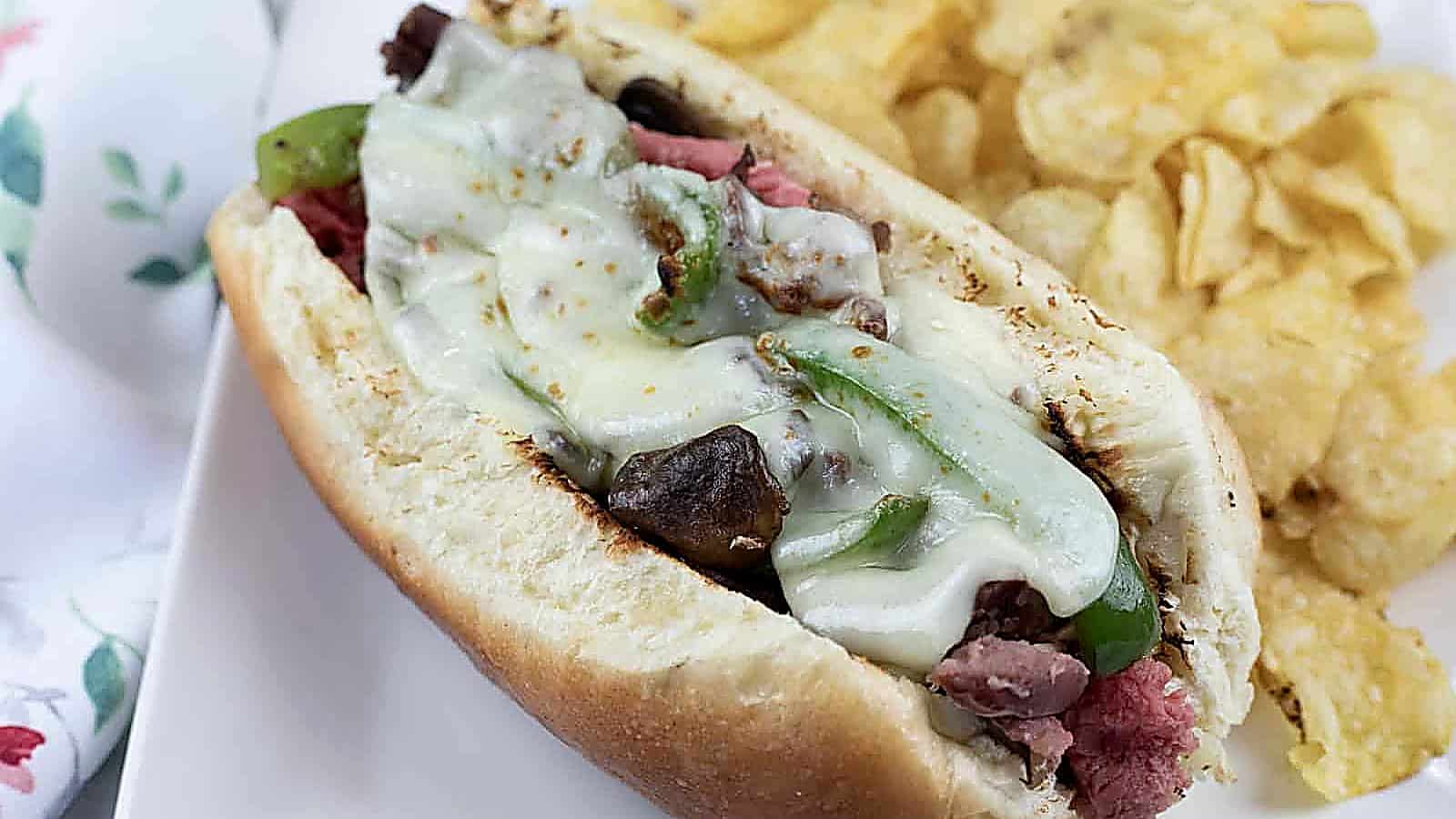 A Philly cheesesteak sandwich with melted cheese, green peppers, and onions in a hoagie roll, served with potato chips on a white plate.