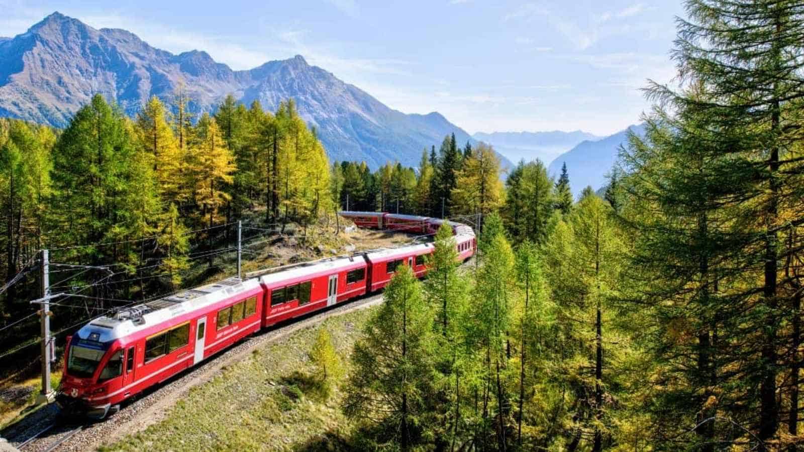 A red passenger train winds through a forested mountain landscape, offering a scenic train travel experience with green trees and distant peaks under a clear sky.