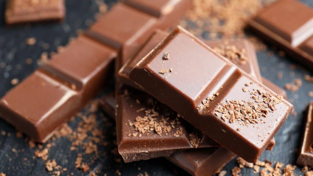Several pieces of milk chocolate bars stacked on a dark surface, with chocolate shavings scattered around them—perfectly capturing the sweet indulgence of National Milk Chocolate Day.