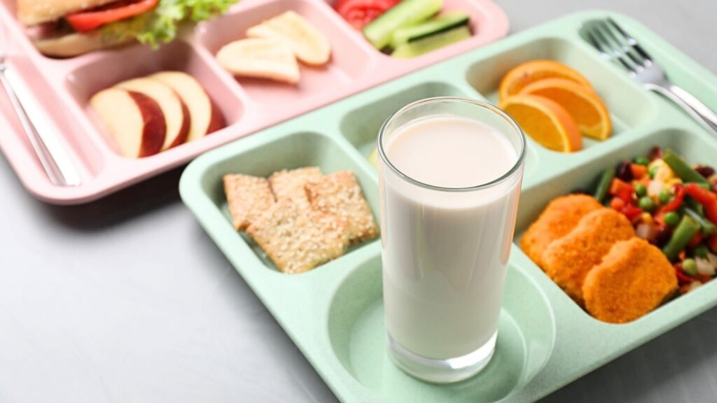 Two compartment trays with a glass of whole milk, assorted fruit slices, crackers, vegetables, and what appears to be breaded patties. Utensils are placed on the trays, reflecting a wholesome and balanced meal option that supports whole milk in schools.