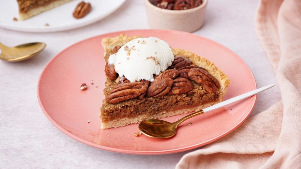 A slice of classic pecan pie topped with a scoop of vanilla ice cream sits on a pink plate with a gold spoon, next to a beige napkin.