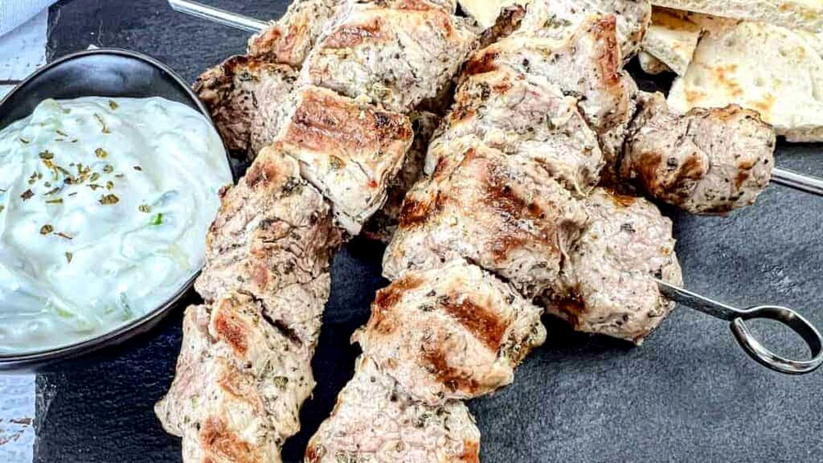 Three pieces of Pork Souvlaki with a bowl of white sauce and pieces of flatbread on a black slate serving board.