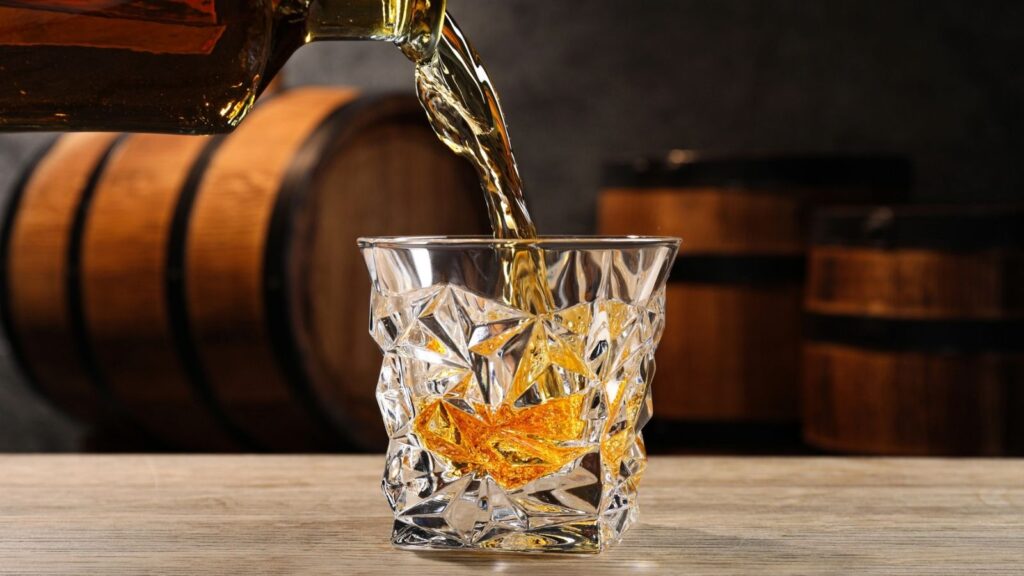 Whiskey is being poured into a faceted glass on a wooden surface, with wooden barrels blurred in the background.