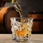 Whiskey is being poured into a faceted glass on a wooden surface, with wooden barrels blurred in the background.