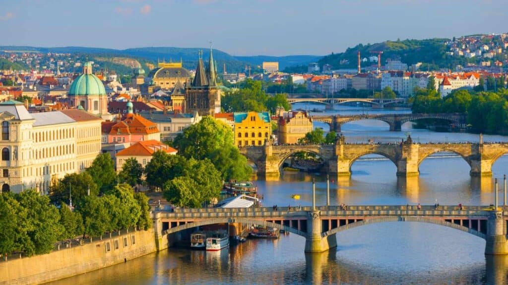 Picturesque view of multiple historic bridges spanning the Vltava River in Prague, with colorful buildings, trees, and hills in the background under daylight.