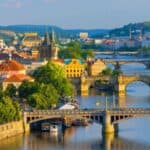 Picturesque view of multiple historic bridges spanning the Vltava River in Prague, with colorful buildings, trees, and hills in the background under daylight.