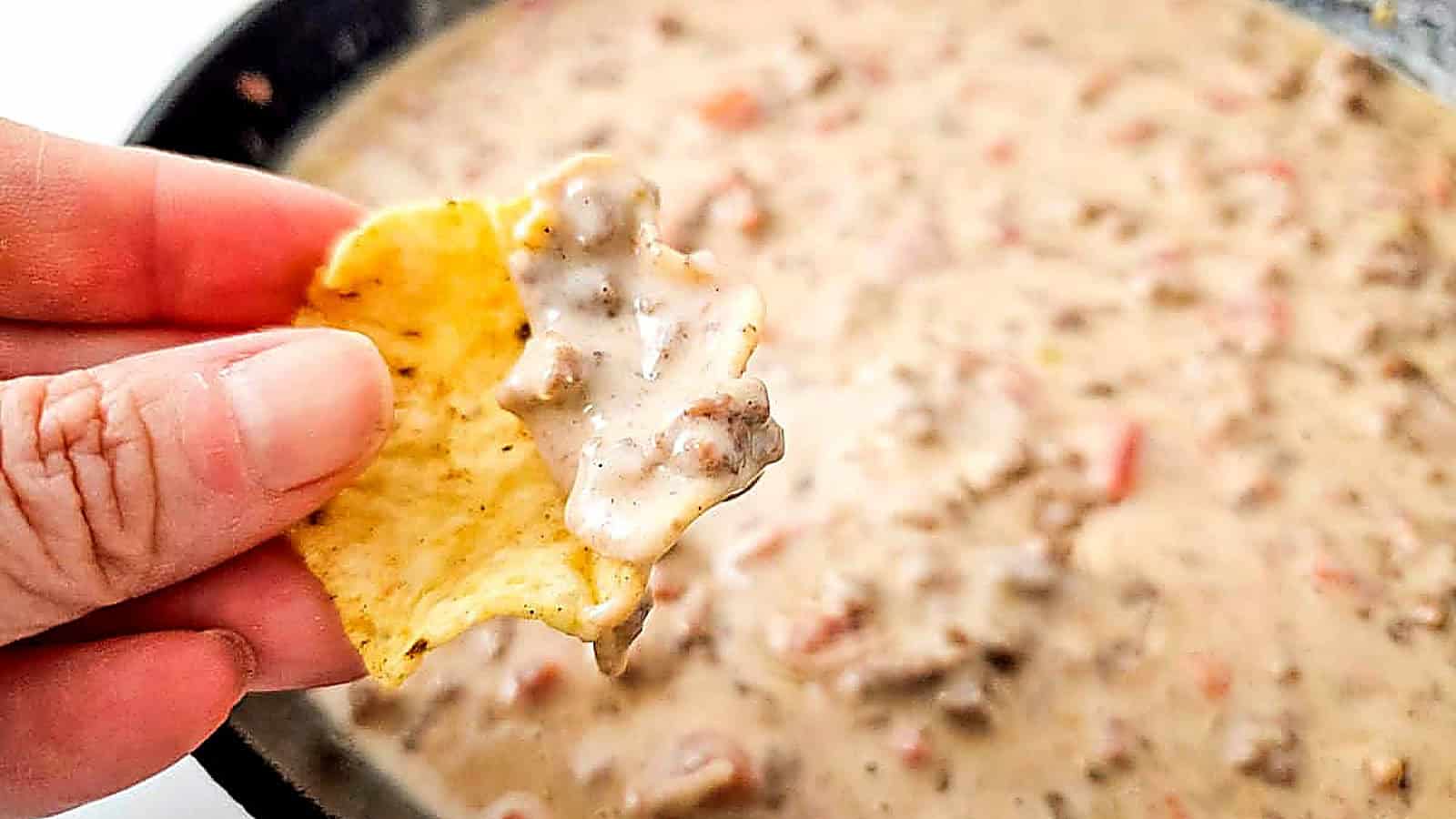 A hand holds a chip dipped in creamy Queso Blanco Rotel Dip over a pan of the same mixture.