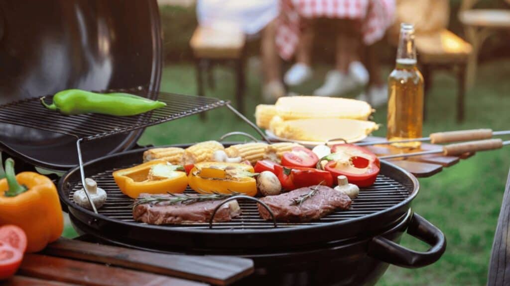 Grill with steaks, corn, sliced peppers, mushrooms, and tomatoes; green pepper above heat; picnic table and beer bottle in background.