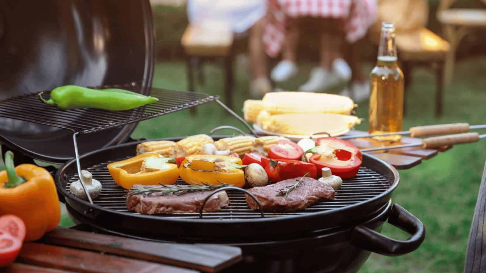 Grill with steaks, corn, sliced peppers, mushrooms, and tomatoes; green pepper above heat; picnic table and beer bottle in background.