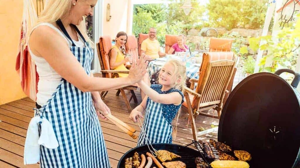 A woman and child in striped aprons high-five near a grill with food, while three people sit at a table outdoors in the background.