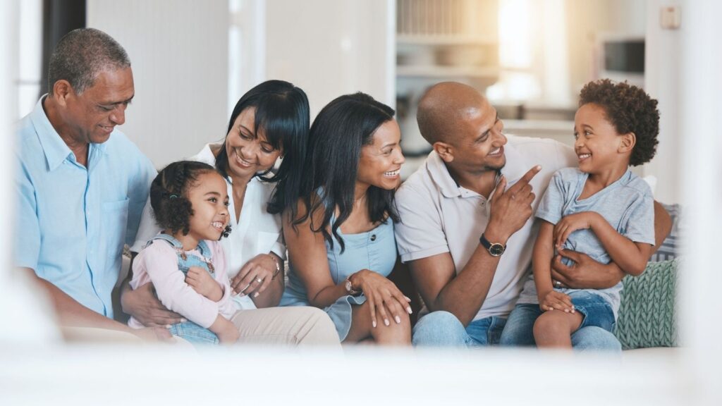 Three adults, two children, and one older adult sit closely on a couch indoors, smiling and interacting warmly with each other.