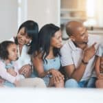 Three adults, two children, and one older adult sit closely on a couch indoors, smiling and interacting warmly with each other.