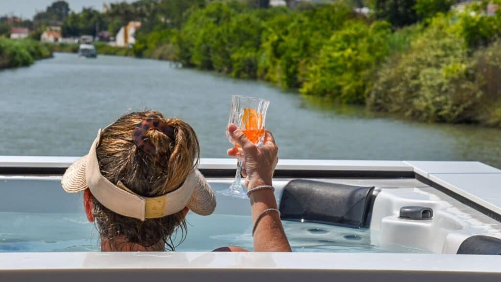 A person in a visor relaxes in a hot tub on a boat, holding a drink, enjoying the tranquil scenery of a southern France barge cruise with a river and lush greenery in the background.