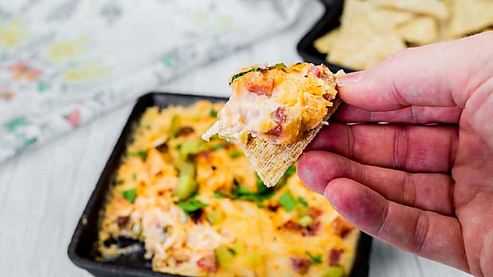 A hand holding a chip topped with cheesy baked dip, with a dish of the same dip and some chips in the background.