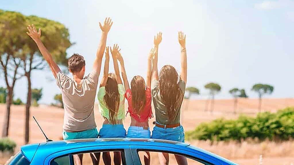 Four people sit on the roof of a blue car with their backs to the camera, raising their arms in the air against a sunny, rural landscape.