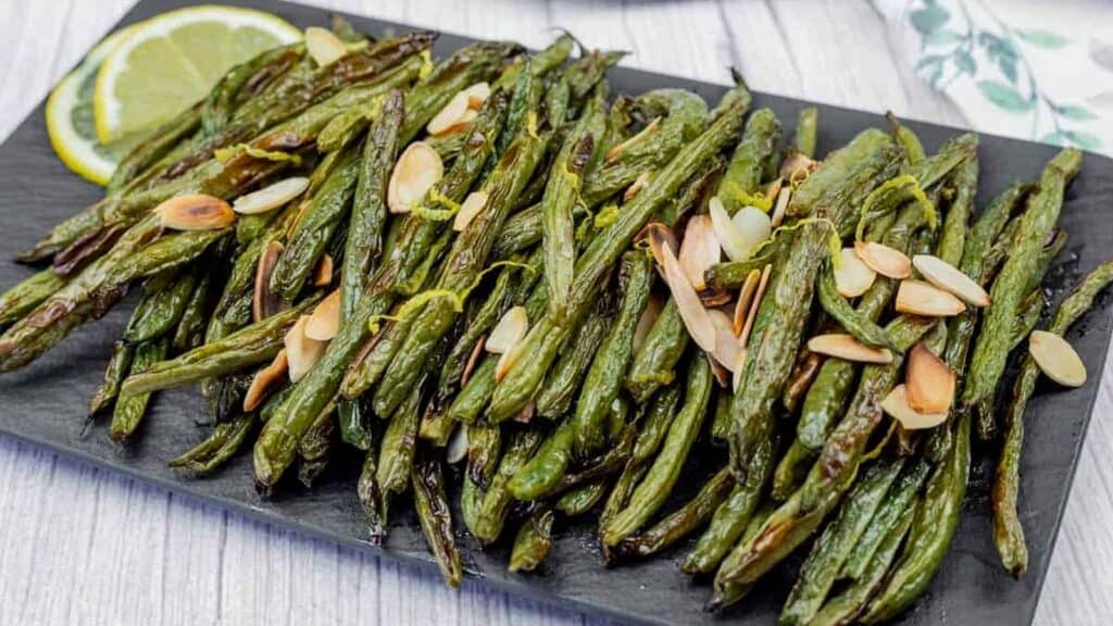 Roasted green beans garnished with sliced almonds, served on a black rectangular tray with lemon slices on the side.