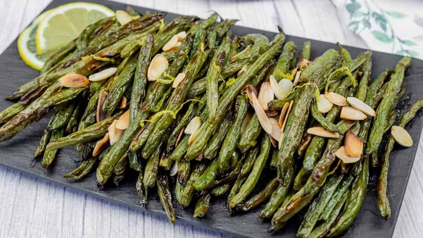 A plate of roasted green beans garnished with sliced almonds, accompanied by lemon slices on a black serving board.