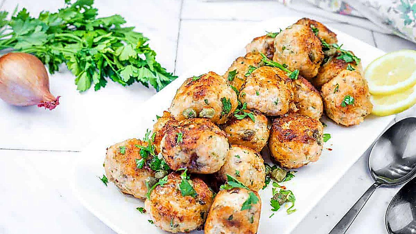 A white plate piled with golden-brown salmon balls garnished with fresh parsley and capers, served with lemon slices.