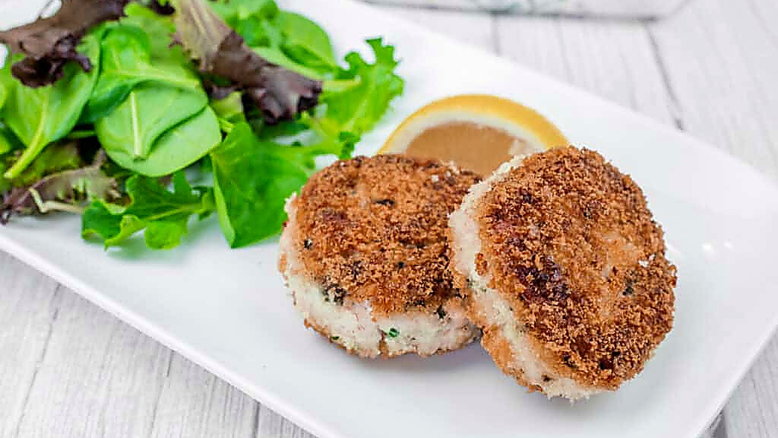 Two Salmon Cakes on a white plate with mixed leafy greens and a lemon wedge.