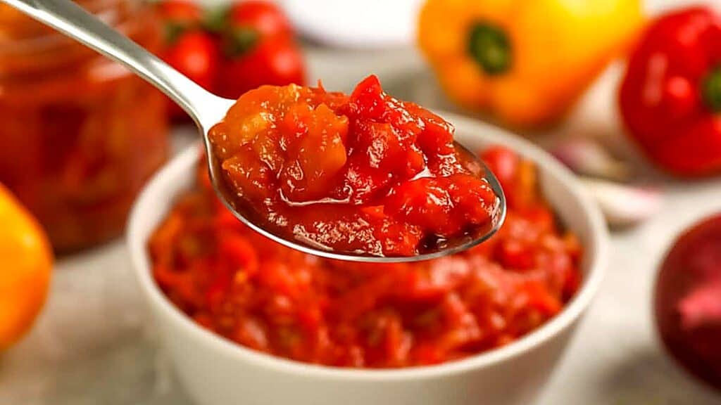A spoonful of chunky tomato salsa is held above a white bowl filled with more salsa; tomatoes and peppers are visible in the background.