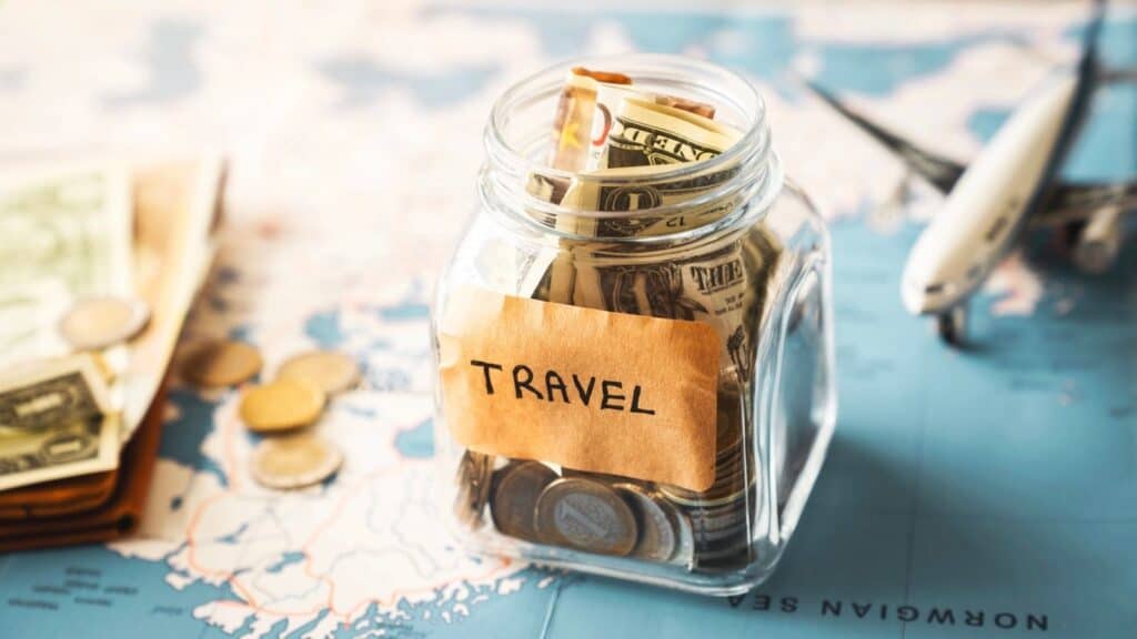 A glass jar labeled "Travel" filled with cash and coins sits on a world map, with a toy airplane and loose money nearby.