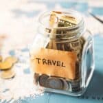 A glass jar labeled "Travel" filled with cash and coins sits on a world map, with a toy airplane and loose money nearby.