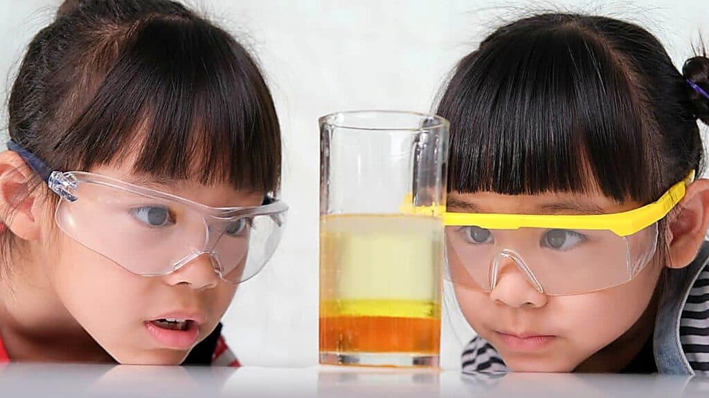 Two children wearing safety goggles observe a glass of liquid with layers of yellow and clear fluid, on a white surface, against a plain background&mdash;an engaging example of science experiments for kids.