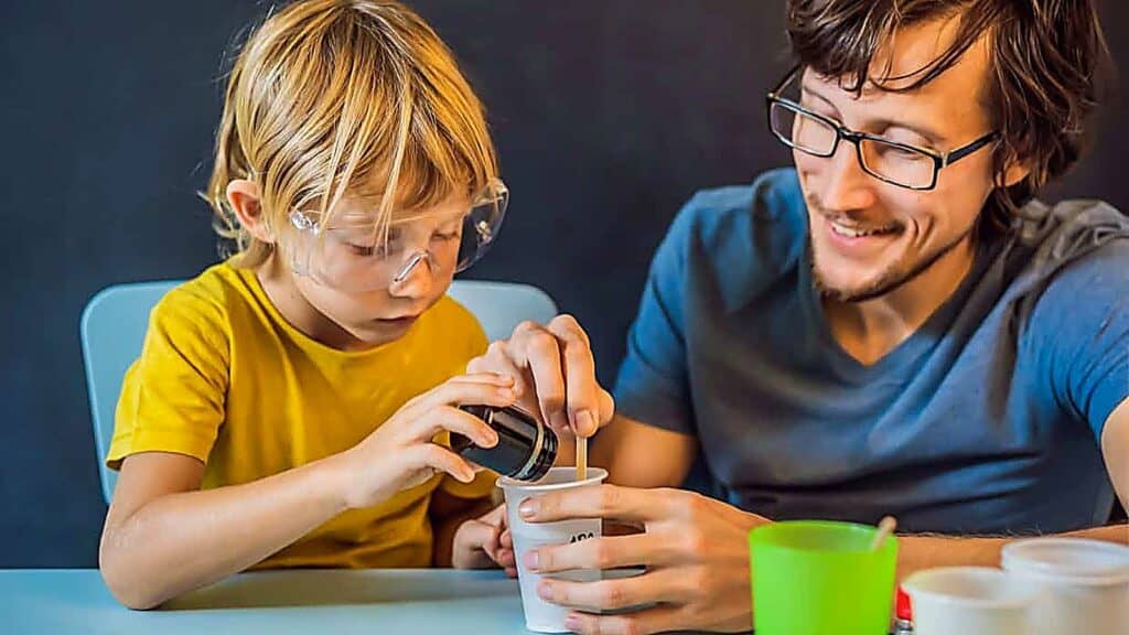 An adult helps a child wearing safety goggles pour liquid into a cup during a science experiment at a table, demonstrating fun and interactive science experiments for kids.