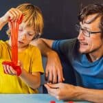 A child wearing safety goggles stretches red slime while an adult sitting next to him smiles and watches; both are seated at a table with craft supplies, enjoying fun science experiments for kids.