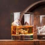 A glass of whiskey with ice cubes sits on a wooden surface, with an empty glass and a wooden barrel in the background.