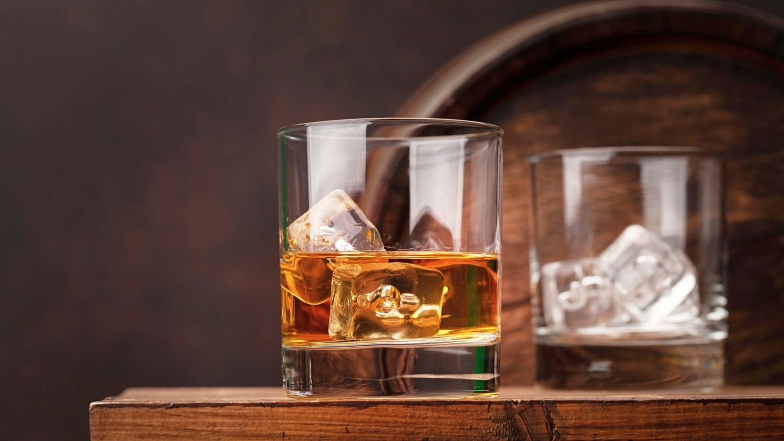 A glass of whiskey with ice cubes sits on a wooden surface, with an empty glass and a wooden barrel in the background.