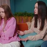 Two young women sit on a sofa; one looks upset with arms crossed, while the other—caught in a subtle power struggle—tries to talk to her, gesturing with her hands.