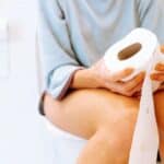 A person sitting on a toilet holding a roll of toilet paper with their pants down in a bathroom.