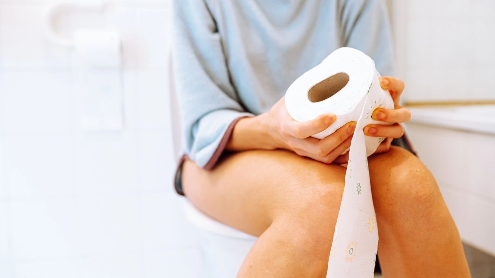 A person sitting on a toilet holding a roll of toilet paper with their pants down in a bathroom.