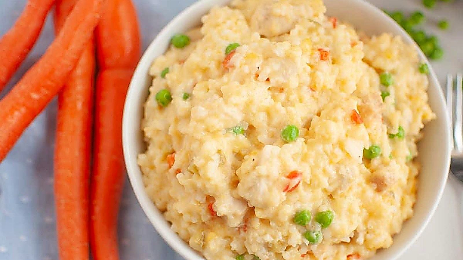 A bowl of Slow Cooker Chicken and Rice is next to whole carrots on a light blue cloth.