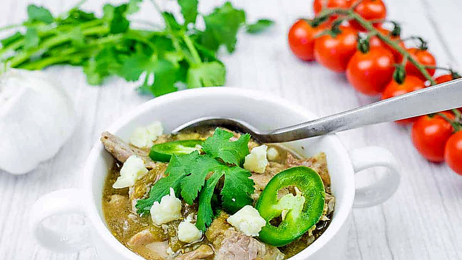 Bowl of Slow Cooker Pork Chile Verde with toppings and a spoon on a white wooden table.
