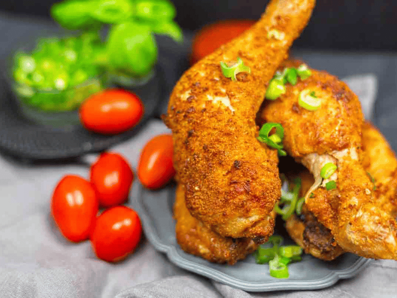 Two crispy fried chicken legs garnished with chopped green onions, served on a plate with cherry tomatoes and green herbs in the background.