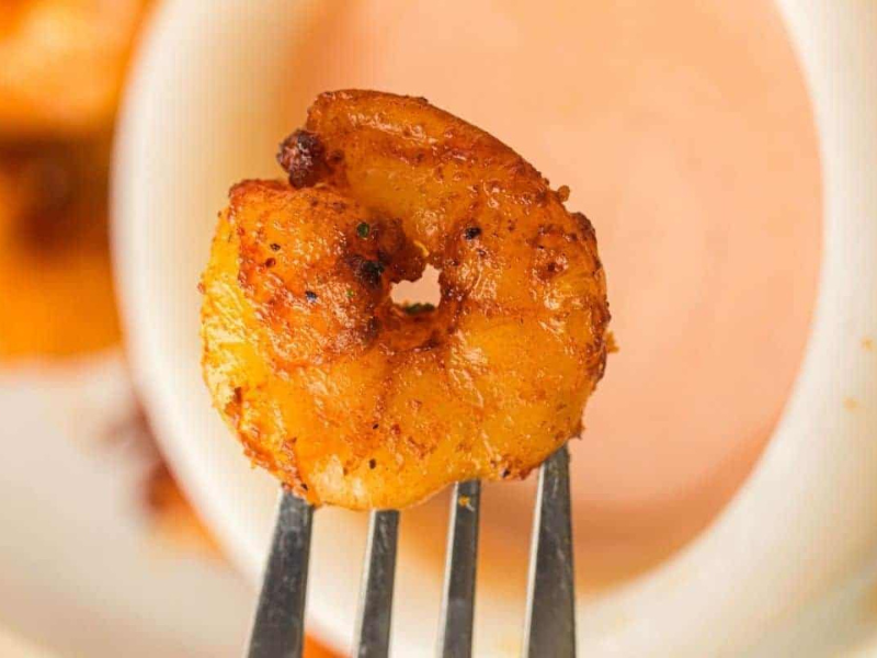A close-up of a seasoned, cooked shrimp on a fork, held in front of a bowl of dipping sauce.