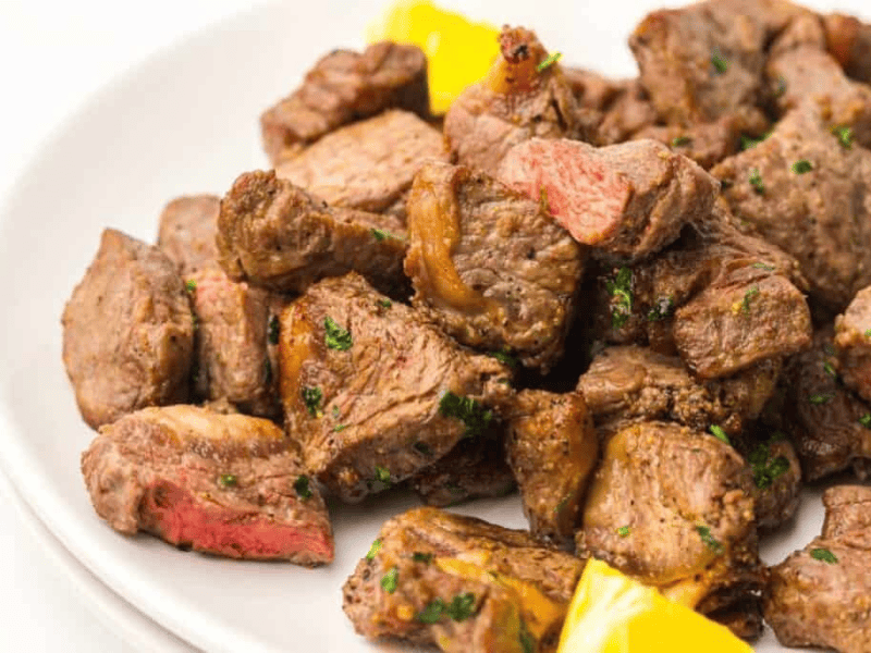A plate of cooked steak bites garnished with herbs, served with pieces of yellow bell pepper.