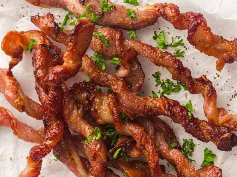 Crispy twisted strips of cooked bacon garnished with chopped parsley, displayed on a white surface.