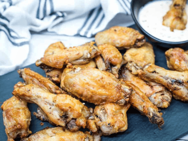 A pile of air-fried chicken wings on a black slate platter with a bowl of creamy dipping sauce in the background.
